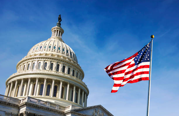 A close-up view of the United States Capitol dome in Washington, D.C., with the American flag waving in the foreground against a bright blue sky.