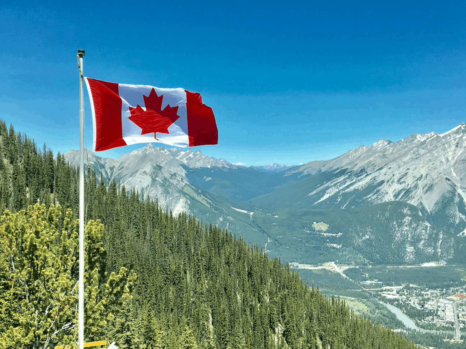 The Canadian flag waving on a flagpole overlooking a vast, mountainous valley with evergreen forests under a clear blue sky.